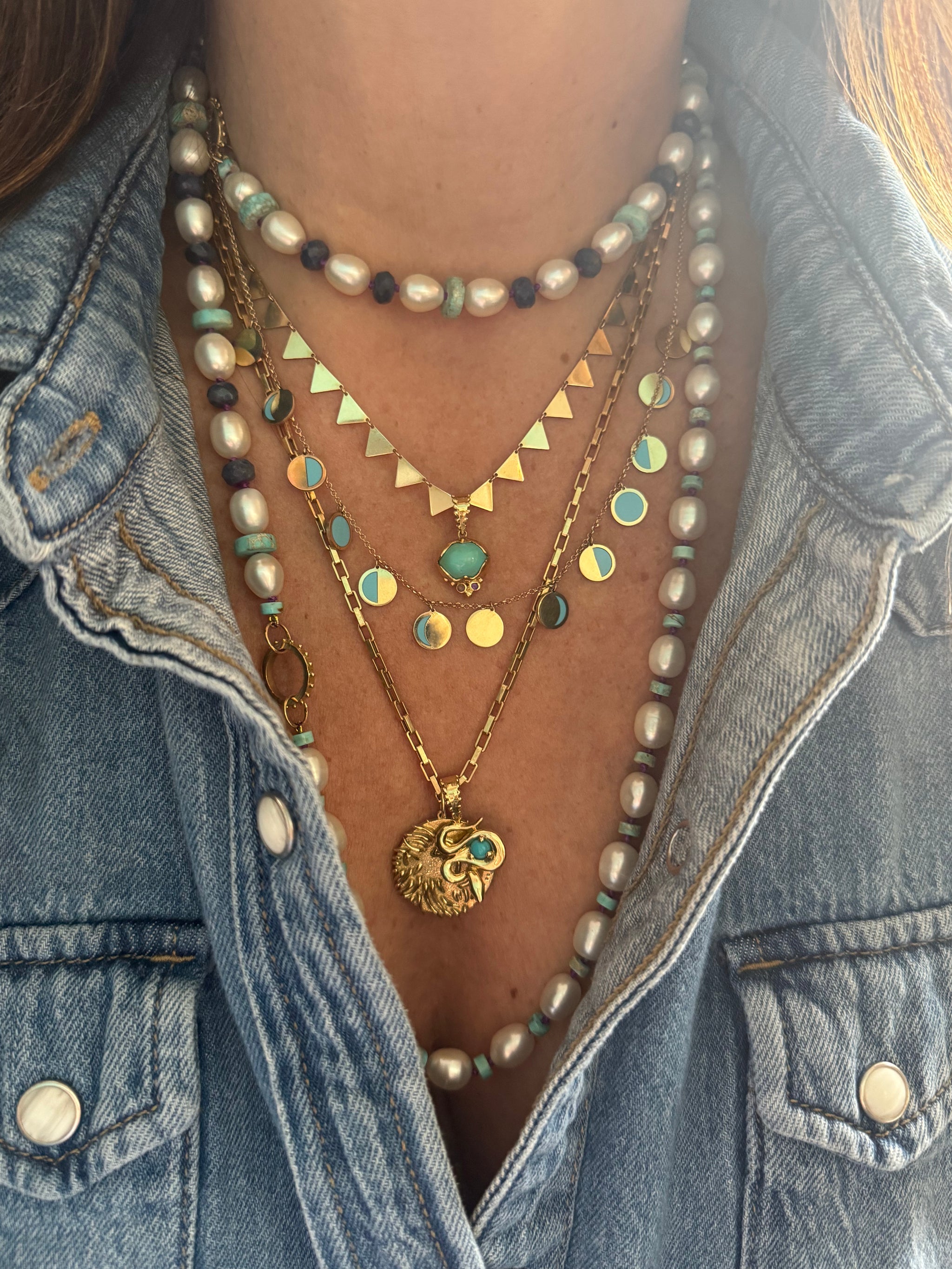 Close-up of pearl strand and gold talisman necklaces with lion, snake, moon phases, lotus, and triangle symbols layered on a model in denim.
